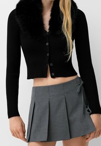 Kvinna iförd en svart ribbad kort cardigan med pälskrage och en grå plisserad mini kjol med rosetter på sidorna, stående mot en enkel bakgrund.