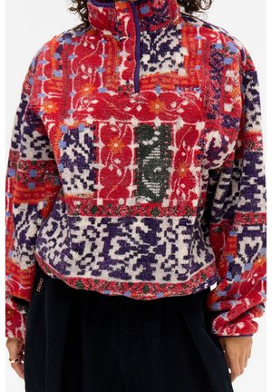Fleece pullover with colorful patchwork floral and abstract patterns in red, purple, orange, and green, worn with black pants.