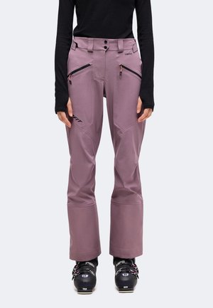 Personne portant un pantalon de ski mauve avec des poches zippées en angle, un haut noir à manches longues et des chaussures de ski noires, de face.