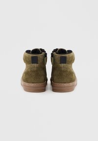 Bottines en daim vert avec une semelle en caoutchouc marron, dotées d'une fermeture éclair et d'une doublure en fourrure noire. Design arrondi avec des accents cousus.