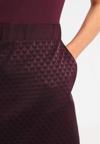 Robe bourgogne avec un haut uni et une jupe texturée à motifs comportant des écailles sombres en répétition. Tissu lisse avec une ceinture en ruban.