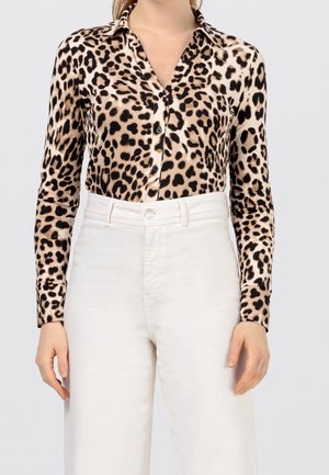 Kobieta ubrana w koszulę na guziki z długim rękawem w cętki, wpuszczoną w białe spodnie z wysokim stanem, na jednolitym tle.