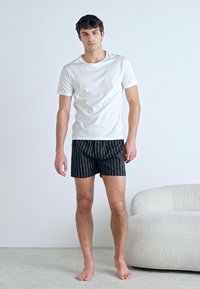 Fehér, rövid ujjú póló fekete csíkos boxeralsóval párosítva. A modell mezítláb áll egy világos színű padlón, a közelben egy textúrált kanapé található.
