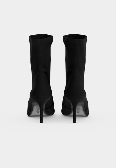Paire de bottines noires à talons vue de dos, avec talons aiguilles pointus et tiges lisses et ajustées sur fond clair uni.
