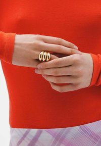 Anillo de oro de tres bandas en una mano, con un fondo de una blusa roja de manga larga. Superficie lisa y brillante con un diseño redondeado y escultural.