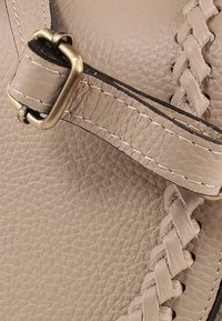 Detail van een beige leren handtas met een gevlochten riem, messing beslag en contrasterende stiksels op een gestructureerd oppervlak.