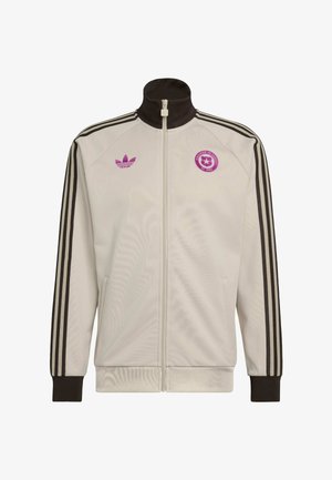 Veste de survêtement Adidas blanche avec fermeture éclair, bandes noires sur les manches, col et poignets noirs, logo Adidas violet et emblème de football du Chili sur la poitrine.
