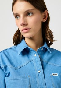 Ljusblå denimskjorta med krage, tryckknappar och två bröstfickor. Har kontrasterande sömmar och en liten logolapp.