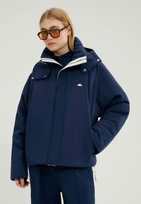 Donkerblauwe pufferjas met een hoge kraag en een afneembare kap. Heeft witte accenten, zakken aan de voorkant en een logo op de borst.