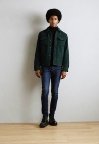Chaqueta de mezclilla texturizada en verde oscuro con dos bolsillos en el pecho, usada sobre un suéter negro de cuello alto; combinada con jeans azul ajustados y botas negras.