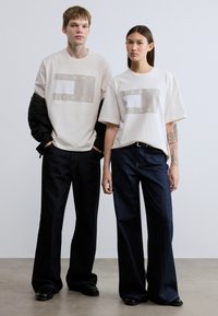 Δύο μοντέλα φορούν υπερμεγέθη, κρεμ μπλουζάκια με γκρι γραφικό σχέδιο που γράφει "TOMMY JEANS", συνδυασμένα με φαρδιά, σκούρα μπλε τζιν παντελόνια.