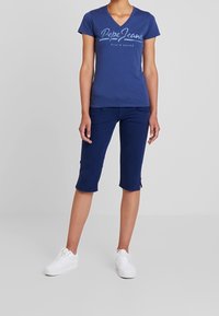 Osoba v modrém tričku s krátkým rukávem ve stylu V (Pepe Jeans), námořnických capri kalhotách a bílých teniskách, stojící na jednolitém pozadí.