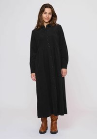 Rochie neagră, cu mâneci lungi și nasturi, dintr-un material moale, având un talie strânsă și o fustă amplă. Este purtată cu cizme până la gleznă, de culoare maro.