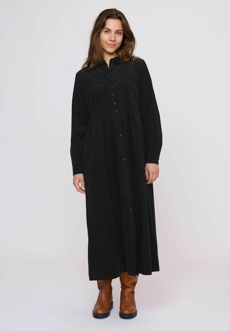 Rochie neagră, cu mâneci lungi și nasturi, dintr-un material moale, având un talie strânsă și o fustă amplă. Este purtată cu cizme până la gleznă, de culoare maro.
