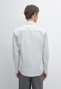 Camicia bianca con righe verticali grigie, vista posteriore. Presenta un colletto standard, maniche lunghe e orlo arrotondato. Il tessuto appare liscio e leggero.