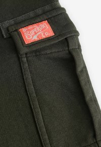Olive green stretch fabric with five-pocket design; features a red fabric label with white text on the back waistband.
