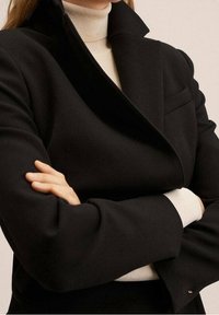 Abrigo negro a medida con un alto cuello y solapas anchas, con una tela texturizada. Capas con un suéter de cuello alto crema debajo.
