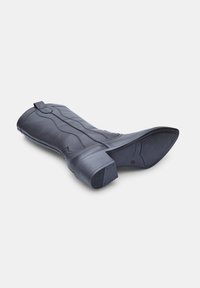 Botín negro de cuero hasta el tobillo con un diseño curvado, superficie texturizada, punta cuadrada y tacón apilado. Presenta una lengüeta y acentos cosidos.