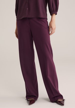 Femme portant un pantalon large violet foncé et un haut boutonné assorti avec manches bouffantes, debout les mains détendues le long du corps.