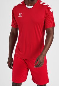 Červený atletický dres s výstřihem do V, krátkými rukávy a bílými akcenty na ramenou; odpovídající červené šortky; vyrobeno z lehkého materiálu.