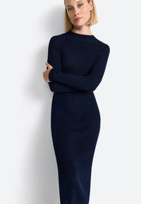 Femme aux cheveux blonds portant une longue robe en maille côtelée bleu marine, les bras croisés contre un fond blanc.