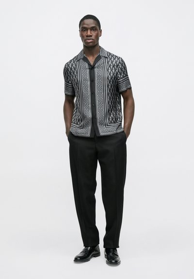 Πουκάμισο με κοντά μανίκια και κουμπιά, με γεωμετρικό ασπρόμαυρο μοτίβο· συνδυασμένο με κομψό μαύρο παντελόνι και γυαλιστερά μαύρα παπούτσια.