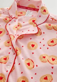 Haut de pyjama rose clair à manches courtes, avec un motif de biscuits et de cœurs, une bordure rouge et une fermeture à boutons. Le tissu est doux et pastel.