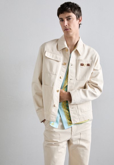 Light beige jacket made of cotton, featuring golden buttons, front pockets, and a logo accent. Worn over a patterned blue shirt.
