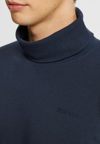 Marineblå turtleneck-tröja i mjukt material, med ribbad krage och präglad logotyp på bröstet, som visar en slät textur.