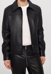Black leather jacket with a classic shirt collar and snap buttons. Smooth texture, fitted design, and distinctive seams across the chest.