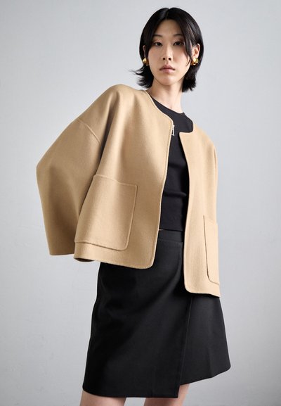 Cappotto in misto lana beige con maniche ampie e tasche frontali, abbinato a un top e una gonna neri. Texture liscia, design minimale.