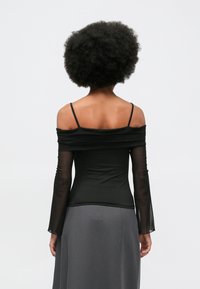 Femeie cu coafură afro, întoarsă, îmbrăcată cu un top negru de tip off-shoulder, cu mâneci lungi transparente, și fustă gri, pe un fundal simplu.