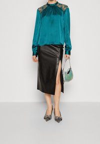 Blouse en satin bleu sarcelle avec détails en dentelle, jupe en cuir noir avec une fente latérale, sac à main argenté brillant, et escarpins noirs pointus avec clous.