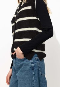 Gilet sans manches en tricot noir avec des rayures blanches, doté d'un col en V, de boutons dorés et d'un ourlet festonné, porté par-dessus un haut noir à manches longues.