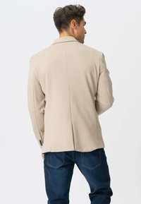 Beige skræddersyet blazer med strukturerede skuldre, enkelt rygslids og subtil glans. Har sorte knapper og en glat stofstruktur.