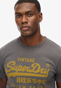 Graues Baumwoll-T-Shirt mit Rundhalsausschnitt, featuring ein großes gelbes bedrucktes Logo "SuperDry" und Text, glatte Textur, lässiges Design.