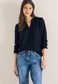 Haut à manches longues bleu marine avec col en V et manches retroussées, associé à un jean bleu clair avec une fermeture à double bouton et une texture délavée.