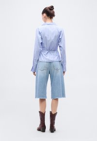 Ανοιχτό μπλε ριγέ πουκάμισο με κουμπιά και εφαρμοστή μέση, συνδυασμένο με ανοιχτόχρωμα τζιν σορτς μέχρι το γόνατο και καφέ μπότες καουμπόι.