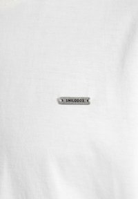 Biała bawełniana koszulka z małą szaro-metalową metką z wytłoczonym napisem "SMILODOX", umieszczoną po lewej stronie na klatce piersiowej. Gładka tekstura.