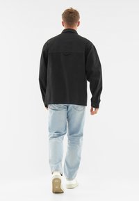 Fekete laza dzseki hátsó zsebfelirattal, világoskék farmerrel és fehér sportcipővel párosítva, háttal látható egy fehér háttér előtt.