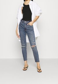 Top negro de tirantes debajo de una camisa blanca de gran tamaño, combinado con jeans azules desgastados y chanclas negras. Presenta detalles rasgados y dobladillo deshilachado.