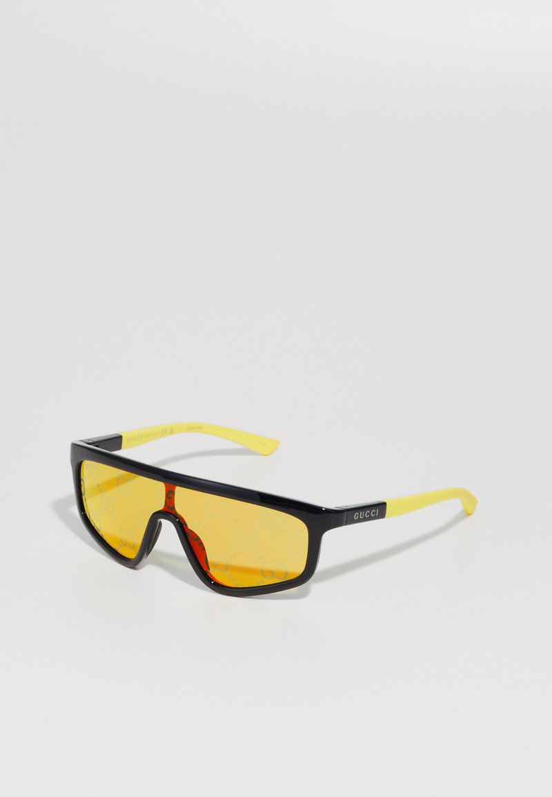 Mustat ja keltaiset aurinkolasit, joissa on rohkea kulmikas muotoilu, keltaiset linssit ja GUCCI-logo mustissa kehyksissä.