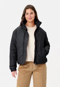 Veste matelassée noire avec col montant, motif matelassé en losanges, fermetures à glissière et à pression, et poignets élastiques, portée par-dessus un haut beige clair.