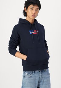 Navy blue hoodie with a front pocket, drawstring hood, and a graphic logo featuring red and blue stripes on the chest. Cotton material.