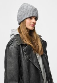 Căciulă tricotată gri cu textură fasonată, purtată de o persoană în jachetă de piele neagră peste un hanorac gri. Cusăturile accentuate sunt vizibile.