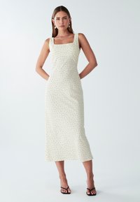 Cremefarbenes Midikleid mit Quadrat-Ausschnitt, schwarzen Punkten und gerader Silhouette. Getragen mit schwarzen Riemchensandalen.