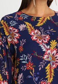 Blouse florale navy avec de grandes fleurs roses et jaunes, des feuilles vertes et une texture de tissu lisse. Encolure arrondie et manches longues.