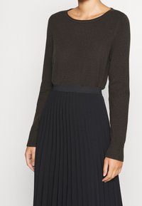 Brauner Langarmpullover mit rundem Halsausschnitt, kombiniert mit einem schwarzen Faltenrock. Der Pullover hat gerippte Bündchen und eine glatte Textur.