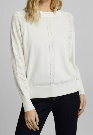 Mujer con un suéter blanco de punto con mangas caladas con patrones y líneas verticales perforadas centrales, combinado con vaqueros oscuros.