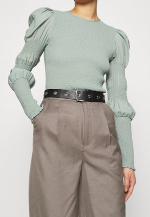 Femme portant un haut smocké vert clair à manches bouffantes, rentrée dans un pantalon taupe taille haute avec une ceinture noire à œillets.
